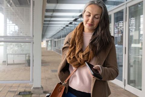 Mature woman checking smartphone in modern glass corridor wearing autumn scarf and coat