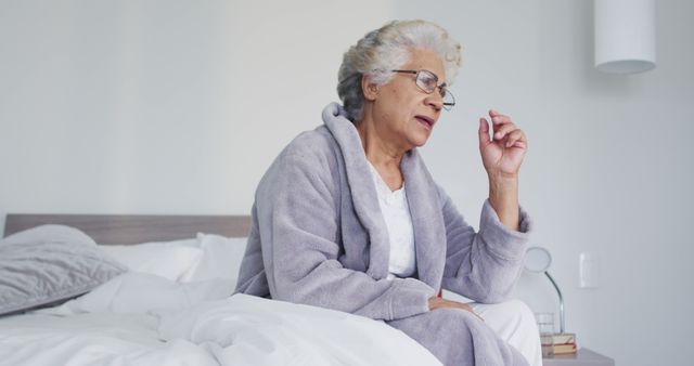 Depicting an elderly woman in a bathrobe, sitting on bed taking daily medication. Ideal for use in healthcare and wellness content, senior care advertising, or articles on daily routines for older adults.
