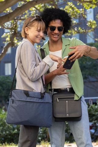 Sunlit urban friends sharing smartphone and laughing while carrying crossbody bags in park