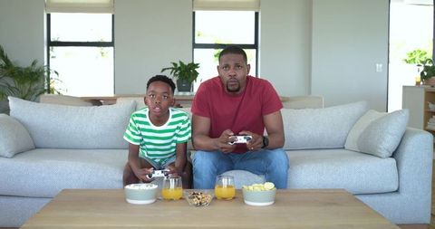 African american father and son playing video games on sofa, bonding over snacks and juice