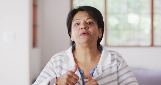 Senior Latina woman gesturing with hands and expressing her thoughts indoors with natural light and soft focus background. Suitable for use in articles about aging, personal stories, communication, and cultural representation.