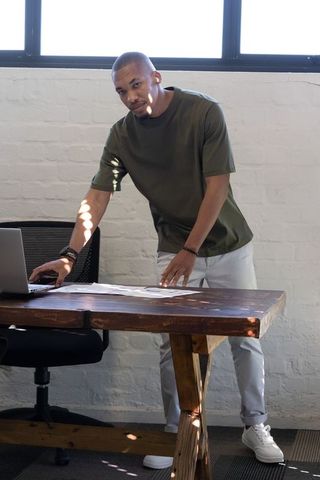 African american man leaning over rustic desk reviewing laptop and papers in bright studio