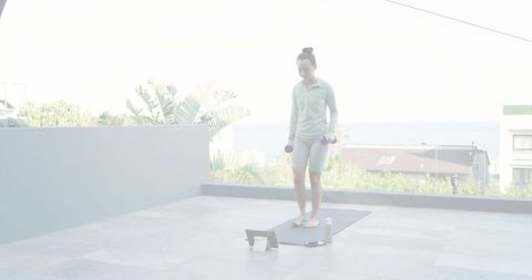 Woman exercising with dumbbells on rooftop yoga mat using tablet overlooking ocean