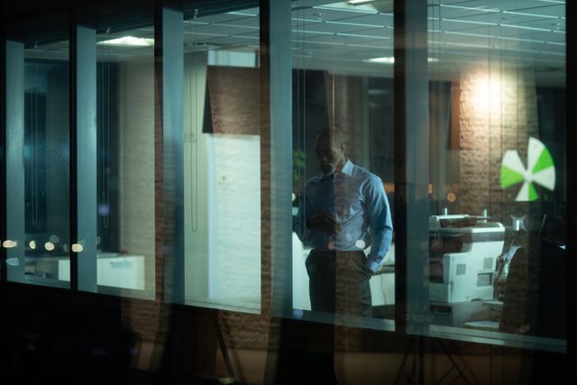 This image depicts an African American businessman working late in a modern office. He is standing and looking through a window, appearing deep in thought. The scene is set in the evening, with the office lights on and reflections visible in the glass. This image can be used to illustrate themes of hard work, dedication, corporate life, and the challenges of balancing work and personal time. It is suitable for articles, blogs, and marketing materials related to business, productivity, and professional life.