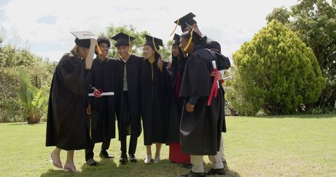 Multicultural graduates celebrating on campus lawn wearing caps and gowns holding diplomas
