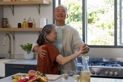 Senior couple dancing in sunlit kitchen by marble island with fresh fruit