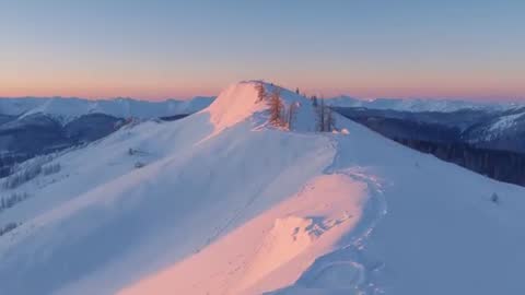 Drone revealing snow-covered alpine ridge at sunrise with footprints and alpenglow
