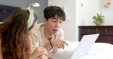 Asian man and african american woman lying on bed browsing laptop in cozy bedroom