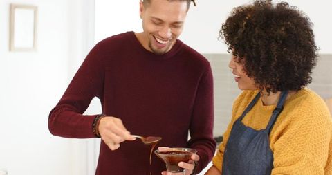 Couple making chocolate sauce in bright kitchen, man spooning dessert while woman watching