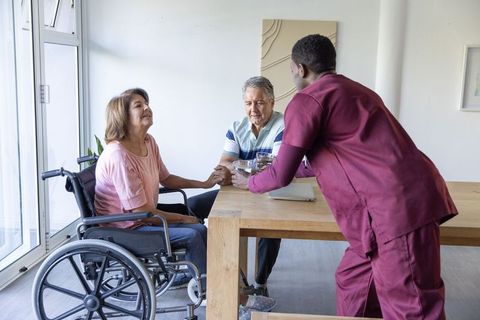 Senior couple sharing tender moment with caregiver offering water in sunlit home interior
