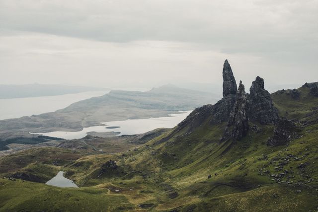 The image captures the iconic Old Man of Storr rock formation on the Isle of Skye in Scotland with surrounding green hills and a lake under an overcast sky. Ideal for use in travel brochures, tourism websites, and nature-inspired content or blogs showcasing rugged landscapes and hiking destinations.