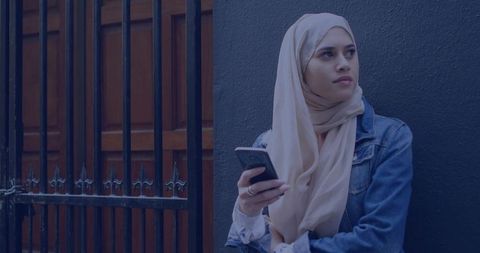 Young woman in beige hijab checking smartphone by metal gate in urban doorway