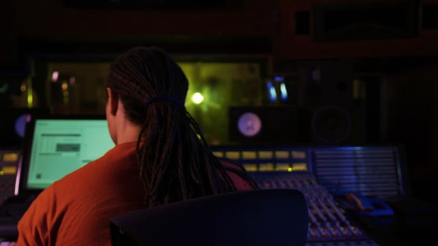 Biracial male sound engineer with long dreadlocks in a ponytail is working at a mixing desk in a dimly lit recording studio. Ideal for use in articles or promotions related to music production, audio engineering, recording studio environment, or professional audio services.