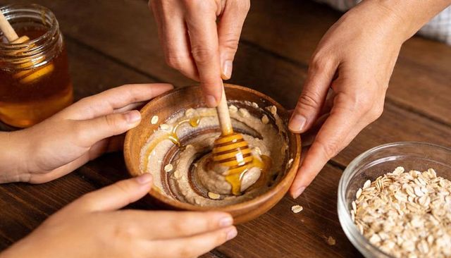 Adult and child stirring honey into oat mixture in wooden bowl on rustic kitchen table