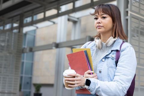 Asian woman standing outside holding notebooks and coffee, wearing denim and headphones