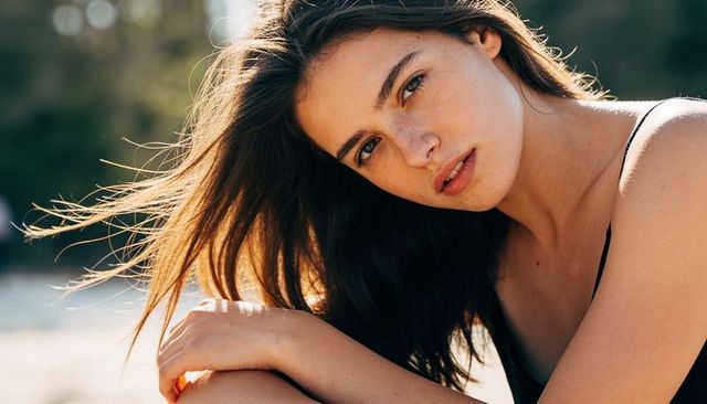 Backlit beach portrait of young woman hugging knee, windblown hair, freckled skin