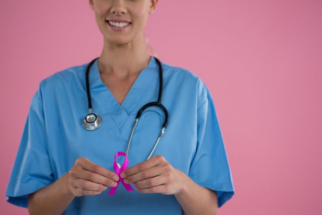 Mid section of happy female doctor in scrubs showing Breast Cancer Awareness ribbon while standing against pink backgrond