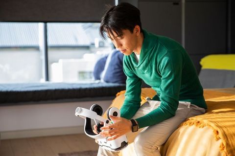 Young asian man examining vr headset on bed in sunlit modern bedroom for home tech setup