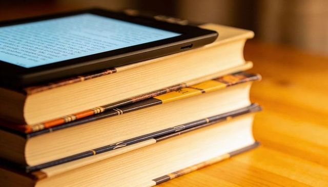 Resting stack of hardcover books with e-reader displaying text on wooden tabletop
