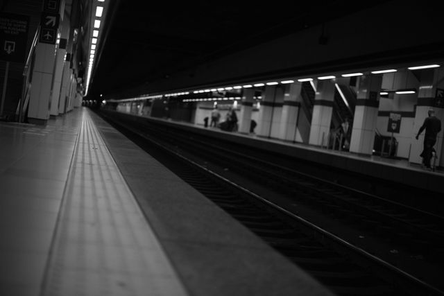 This image features an empty train station platform at night, captured in black and white. The modern rail tracks and dim lighting evoke a sense of solitude and quiet urban life. It is suitable for use in travel-related content, city life documentaries, transportation industry visuals, or artistic projects focusing on modern solitude and perspective.