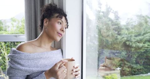 Pensive midlife woman leaning by window holding mug wearing cozy grey off-shoulder sweater