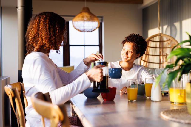 Lesbian couple enjoying a relaxed breakfast together in a bright, sunny kitchen. One is pouring coffee while the other smiles warmly. Ideal for use in articles or advertisements focusing on LGBTQ+ relationships, morning routines, domestic life, and happiness.