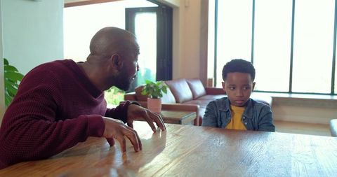 African american father leaning toward son at wooden table expressing concern and tension