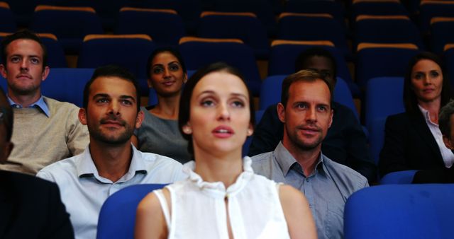 A diverse group of young and middle-aged adults is attentively watching a presentation or event in a theater with blue seats, with copy space. Their focused expressions suggest engagement with the content being presented.
