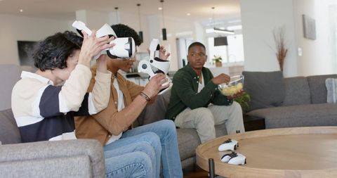 Multicultural friends playing VR games on modern couch with controllers and snacks