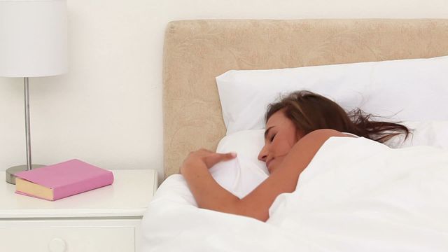 Brunette woman reaches out from under the covers to turn off her alarm in a cozy bedroom. Perfect video for topics such as morning routines, restful sleep, lifestyle, and home relaxation. Can be used in articles, advertisements, blog posts, and social media related to sleep habits, productivity, home decor, and wellness.