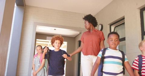 Teacher guiding diverse elementary students walking along school porch with backpacks