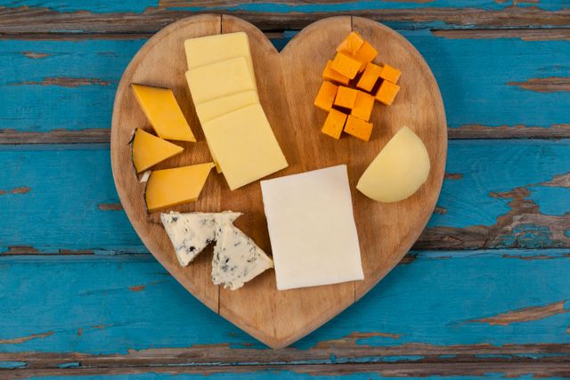 Assorted cheeses displayed on a heart shaped wooden board against a rustic blue background. Ideal for use in culinary blogs, gourmet food promotions, dairy product advertisements, or social media posts about food and cooking.