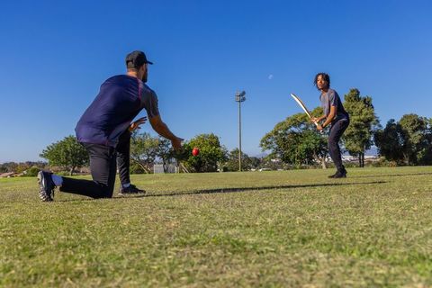 Diverse men training cricket in park kneeling coach releasing ball batter ready to strike