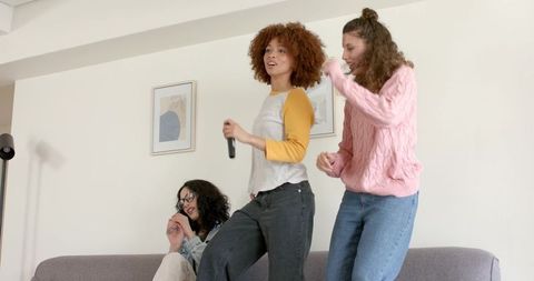 Multiracial friends singing and dancing on couch during home karaoke party, casual fun