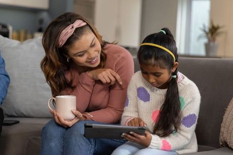 Diverse mother smiling while daughter using tablet and holding mug on cozy sofa at home