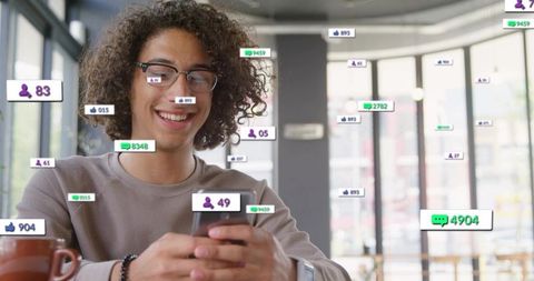 Smiling young man with glasses in cafe scrolling smartphone with social reactions overlays