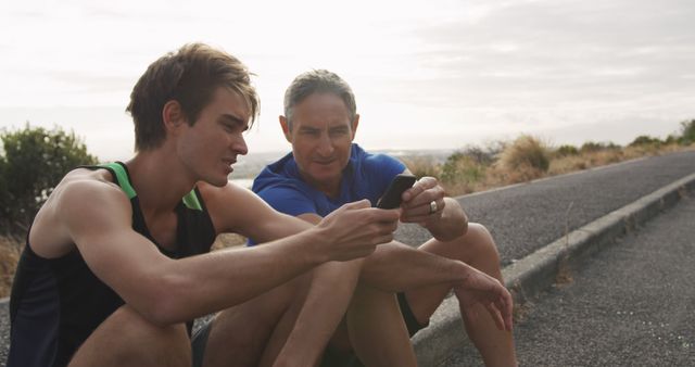 Mature man and young man sitting side-by-side on a pavement outdoors, both dressed in casual athletic wear. They are looking at a smartphone, discussing something together. This image can be used to illustrate family bonding, fitness routines, technology use in exercise, or father-son relationships.