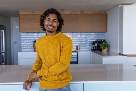 Indian man in mustard sweater leaning and smiling on kitchen island in modern home