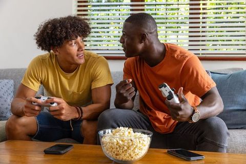 Two friends playing console games on couch with controllers, popcorn, casual competition