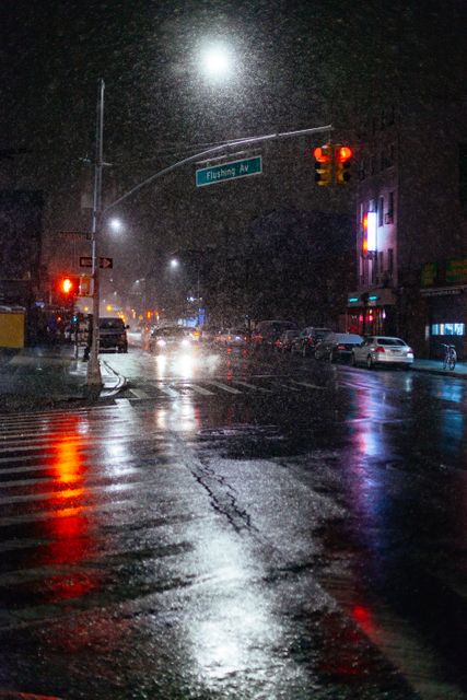 Rainy nighttime city street scene with reflections from streetlights and car headlights on wet pavement. Street sign for Flushing Avenue visible. Atmosphere reflects urban hustle in inclement weather conditions. Useful for illustrating urban life, nighttime transportation, city traffic, and rainy weather effects.