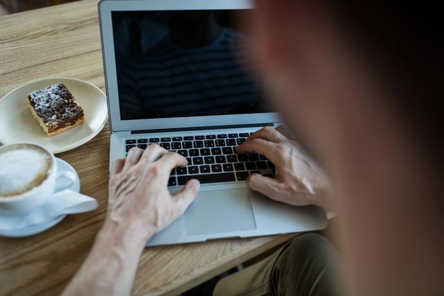 Man typing on laptop in coffee shop, with coffee and pastry on table. Ideal for illustrating remote work, freelance lifestyle, productivity, and casual work environments.