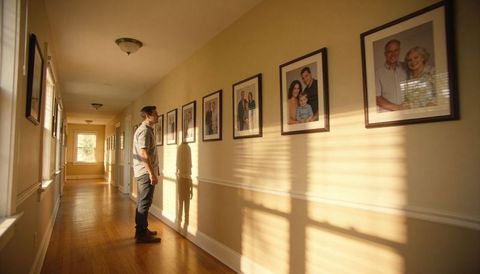 Man standing studying framed family portraits in warm sunlit hallway with hardwood floors