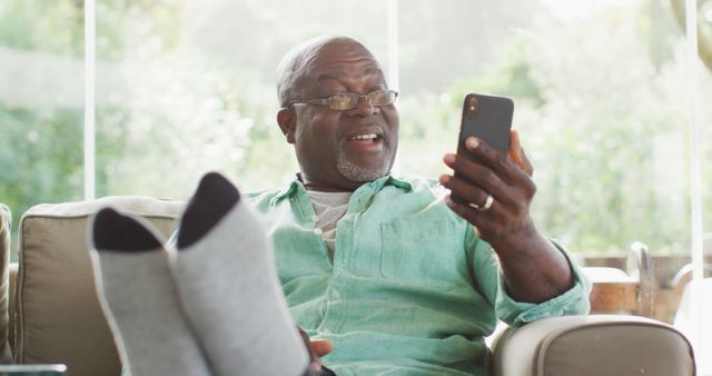 Senior man sitting on a couch in a living room, wearing glasses and a casual green shirt while talking and smiling at his smartphone. Ideal for usage in lifestyle articles, technology advertisements, senior living blog posts, or communication services promotions.
