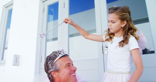 Girl holding magic wand pretending to crown man; girl dressed in fairy costume while man wears tiara; playful interaction; outdoor family fun; concept of imagination and bonding; suitable for parenting, lifestyle, and family-related content.