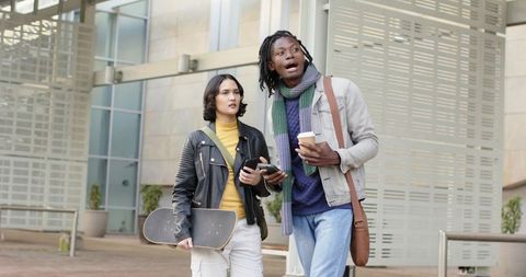 Two young friends walking across campus carrying skateboard and coffee while checking phone