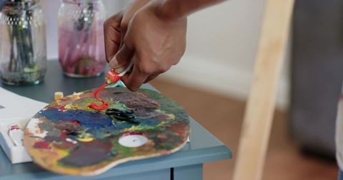Artist squeezing red paint onto wooden palette on blue side table, studio closeup