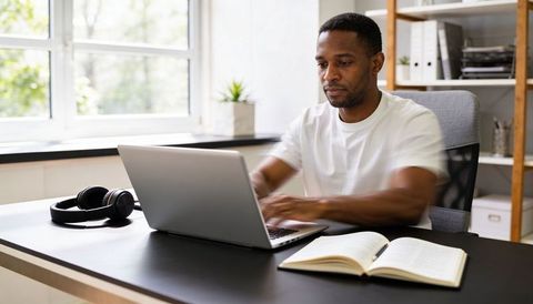 Typing remote professional at minimalist home office with laptop, open notebook, headphones