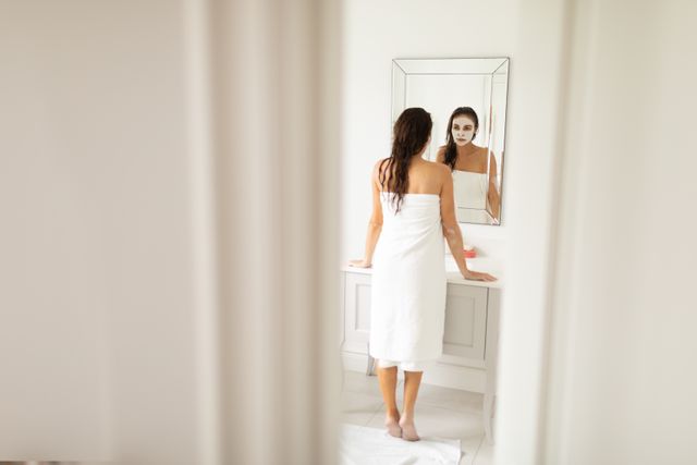 Rear view of woman in wrapped towel and facial mask standing in front of mirror at bathroom
