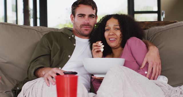 Image depicts a couple in home setting. They are sitting on a couch wrapped in blankets, sharing a bowl of popcorn while watching TV. Ideal for concepts related to home entertainment, quality time, relaxation, and cozy moments. Suitable for use in advertisements, home living blogs, and lifestyle articles.
