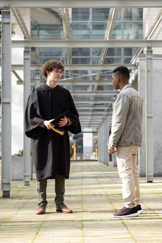Graduate in gown holding mortarboard talking with friend beneath modern glass canopy